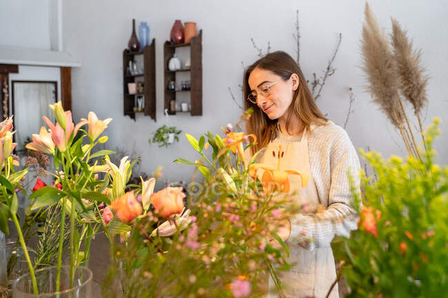 Florista femenina joven concentrada en delantal y anteojos que arregla flores amarillas fragantes en jarrón mientras trabaja en una tienda de flores - foto de stock