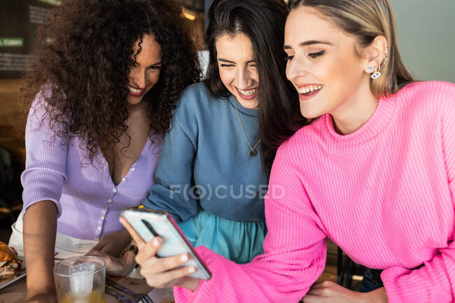 Sonrientes amigas jóvenes que usan ropa casual navegando por teléfonos móviles mientras se reúnen para almorzar en el restaurante - foto de stock
