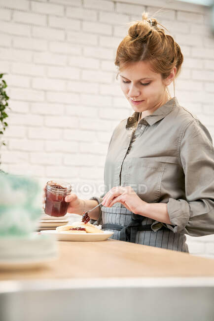 Jovem fêmea com jarra colocando deliciosa geléia de baga em panquecas enquanto cozinha na mesa na cozinha — Fotografia de Stock