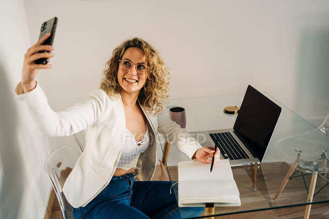 From above of cheerful young female freelancer freelancer with curly hair in stylish outfit taking selfie on smartphone while working remotely on project using laptop at home — Foto stock