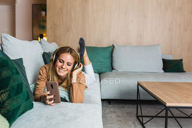 Радісна жінка лежить на дивані і насолоджується музикою в навушниках, дивлячись на екран смартфона — стокове фото