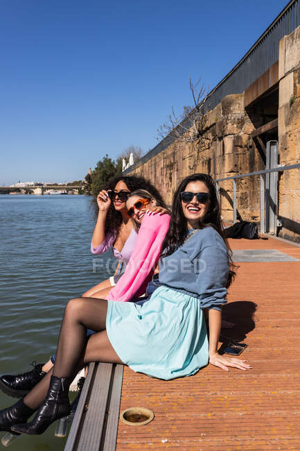Fröhliche multiethnische Freundinnen in lässigen Outfits sitzen am Ufer der Stadt und schauen lächelnd in die Kamera an einem sonnigen Sommertag — Stockfoto