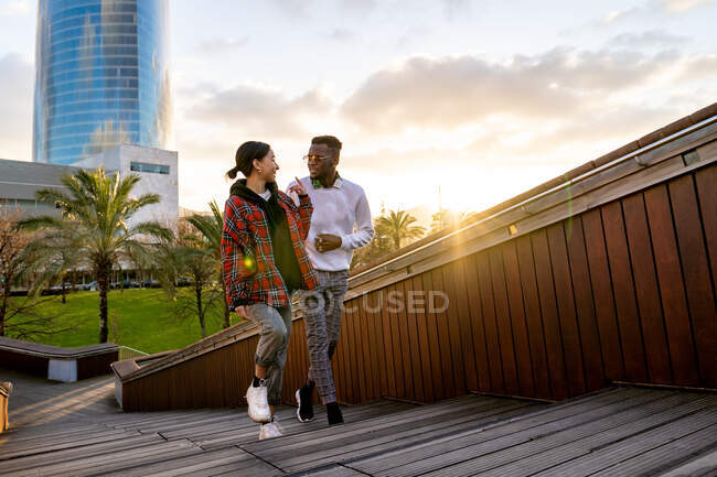 Веселая многонациональная пара в стильной одежде, говорящая, глядя друг на друга и прогуливаясь по городской лестнице под солнечным светом — стоковое фото