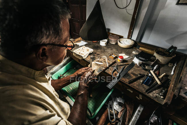 Зверху концентровані етнічні чоловіки похилого віку в повсякденному одязі і окуляри загострюють інструменти під час роботи за дерев'яним столом у традиційній майстерні. — стокове фото