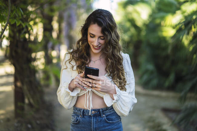 Привлекательное содержание женщины в повседневной одежде с обнаженным животом просматривает мобильный телефон в обильном парке в ясную теплую погоду — стоковое фото