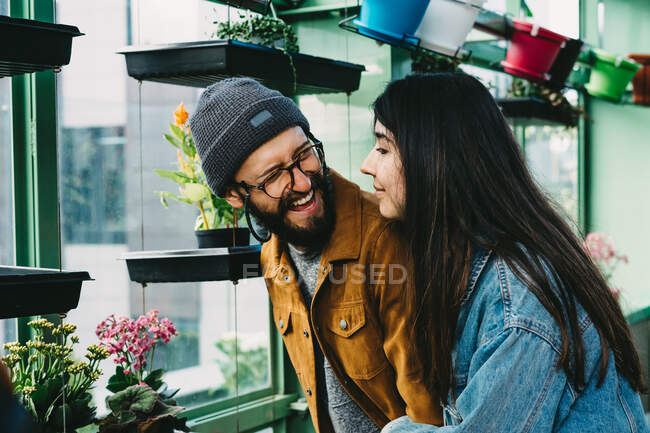 Content stylish couple embracing in greenhouse while standing near shelf and picking blooming potted flower — Stock Photo