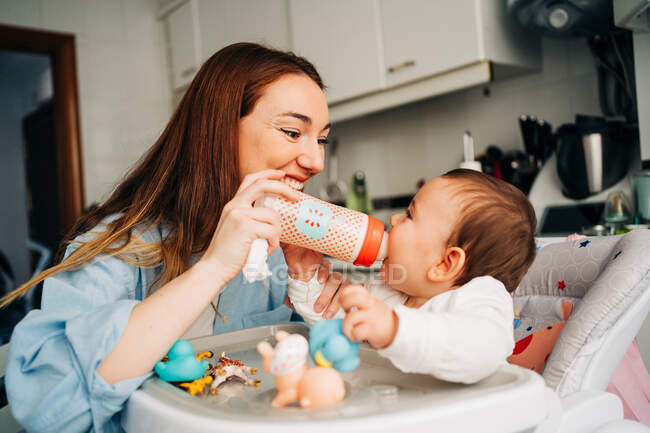 Fröhliche junge Mutter mit Babyflasche in lässigem Outfit füttert entzückendes kleines Baby im Hochstuhl in moderner Küche — Stockfoto