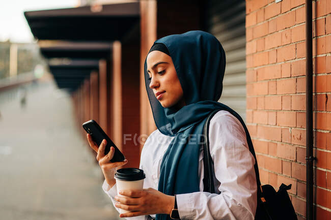 Side view of ethnic female in hijab standing on platform on railway station and browsing mobile phone while waiting for train — Stock Photo