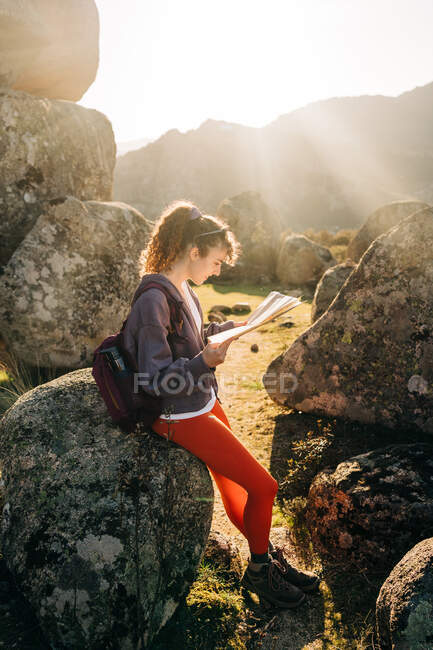 Вид сбоку счастливой молодой женщины с темными волосами в повседневной одежде, сидящей среди скальных валунов и читающей карту на фоне безоблачного голубого неба — стоковое фото