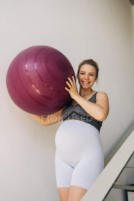Jovem sorrindo expectante fêmea em roupas esportivas com bola de ioga passeando em escadaria em casa — Fotografia de Stock