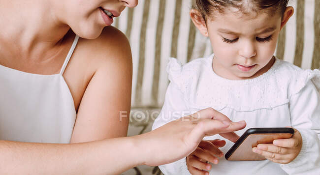 Mulher de cultura ajudando a filha pequena étnica com o telefone móvel de navegação enquanto sentado no sofá em casa — Fotografia de Stock