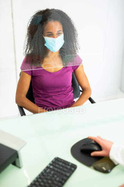 African American woman in face mask during appointment sitting at cropped unrecognizable doctor office desk protected by a perspex glass shield screen at modern clinic during coronavirus outbreak — Stock Photo