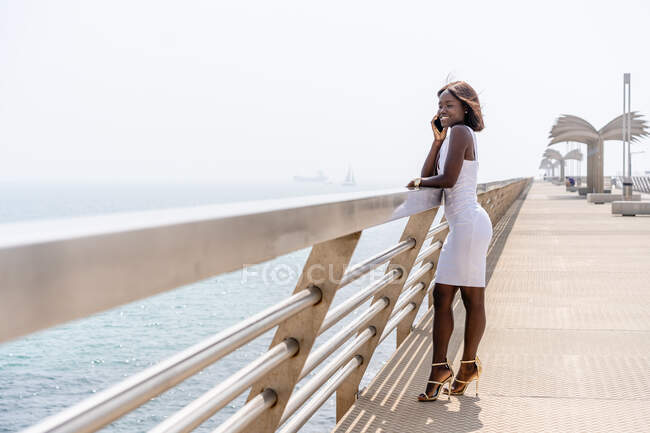 Seitliche Ansicht in voller Länge hinreißende Afroamerikanerin in trendigem weißen Kleid, die bei sonnigem Sommerwetter in der Nähe der Stadtpromenade steht und sich per Smartphone unterhält — Stockfoto