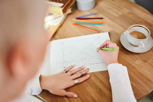Зверху врожаю невизначена жінка-астролог робить нотатки на папері з геометричним малюнком за столом з чашкою кави — стокове фото