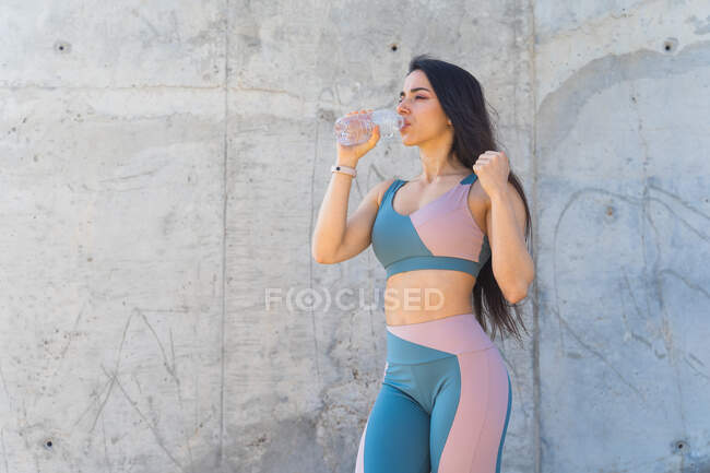 Thirsty female athlete in sports clothes drinking water from bottle after working out while looking away on gray background — Stock Photo