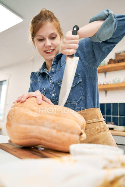 Jovem fêmea com faca afiada cortando abóbora crua na tábua de corte enquanto cozinha na cozinha em casa — Fotografia de Stock