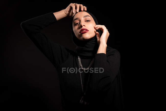 Приваблива молода ісламська жінка, одягнена в чорне вбрання і хіджаб торкається обличчя м'яко і дивиться на камеру — стокове фото