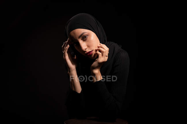 Attraente giovane donna islamica vestita di nero e hijab che tocca delicatamente il viso e guarda la fotocamera — Foto stock