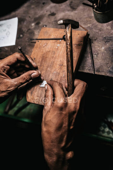 From above faceless craftsman holding metal piercing saw and piece of silver while working at shabby desk — Stock Photo