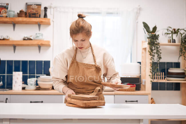 Jovem focado fêmea em avental com sortidas tábuas de corte de madeira e placa à mesa em casa de luz — Fotografia de Stock