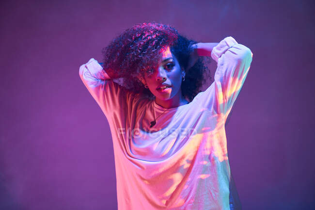 Portrait of young African American female dancer in loose informal wear touching curly hair and looking at camera while dancing in dark studio in neon lights — Stock Photo