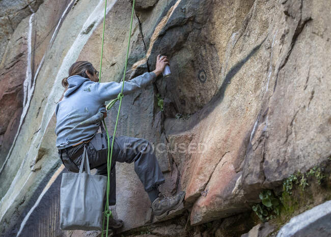 Vista laterale corpo pieno di pittore con vernice spray facendo graffiti appesi sulla corda su ripido pendio roccioso — Foto stock