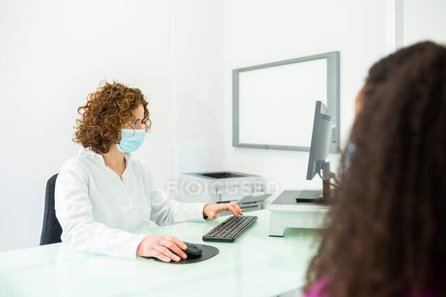 Recadrée femme afro-américaine méconnaissable pendant le rendez-vous assis chez la femme médecin dans le masque facial avec bureau protégé par un écran en verre perspex à la clinique moderne pendant l'épidémie de coronavirus — Photo de stock