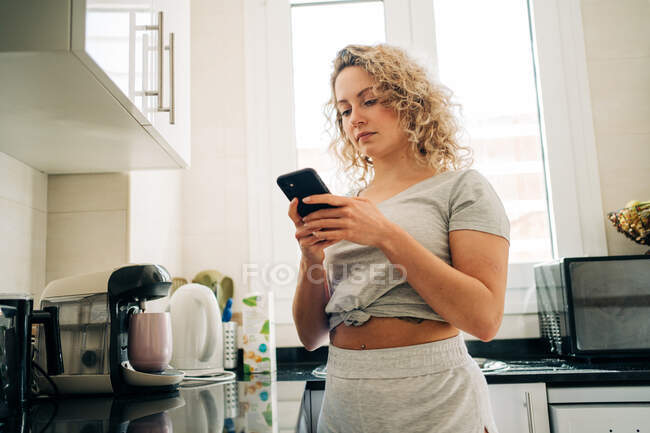 Неэмоциональная молодая женщина в повседневной домашней одежде просматривает мобильный телефон, стоя на современной кухне — стоковое фото