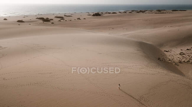 Зверху анонімної жінки-туристки в білій сукні, що йде на ребристий пісок під світлим небом — стокове фото