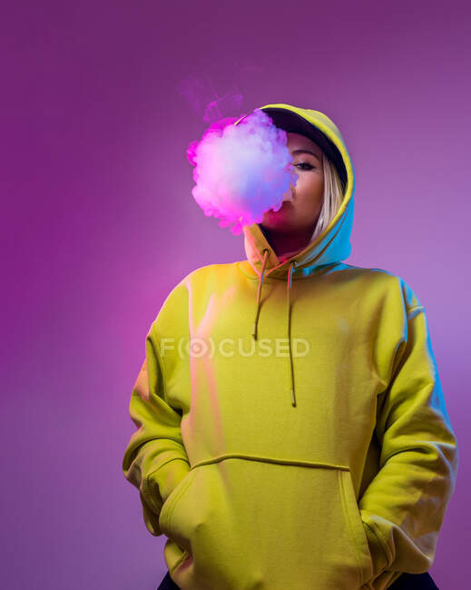 Confiado hipster femenino en sudadera con capucha fumar cigarrillo electrónico en el estudio sobre fondo rosa y mirando a la cámara - foto de stock