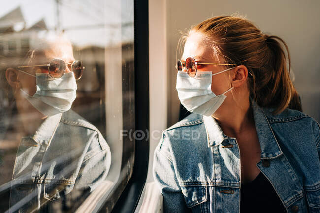 Молода жінка в джинсовій куртці і захисна маска для обличчя дивиться у вікно поїзда під час роботи — стокове фото