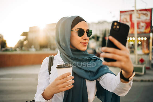 Femme ethnique en foulard et lunettes de soleil élégantes debout avec boisson à emporter dans la rue et prendre selfie avec téléphone portable — Photo de stock