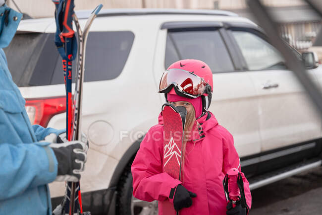 Nettes kleines Mädchen in rosa warmer Sportbekleidung versteckt Gesicht hinter Skiern, während es auf Skiclub-Parkplatz steht und zufrieden in die Kamera schaut — Stockfoto