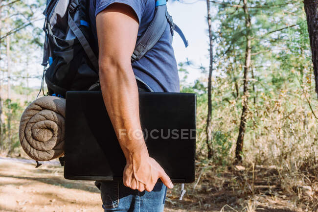 Seitenansicht der Ernte unkenntlich männlichen Nomaden mit Habseligkeiten stehen auf Fußweg mit Netbook in sonnigen Wäldern — Stockfoto