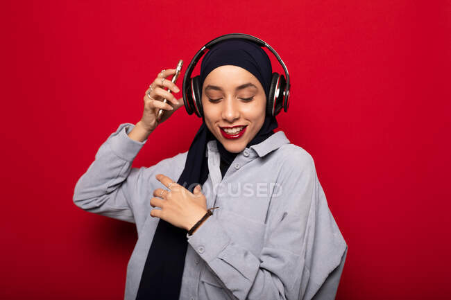Smiling attractive Muslim female in casual clothes and hijab listening to favorite music via headphones on smartphone dancing against red background in studio looking down — Stock Photo