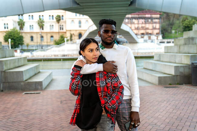 Moda afro-americano masculino abraçando etnia feminina amado com mão no bolso no pavimento urbano — Fotografia de Stock