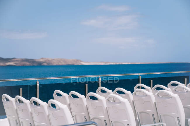 Leere weiße Stühle an Deck eines Kreuzfahrtschiffes, das in blauem Meerwasser mit Berg an Land segelt — Stockfoto