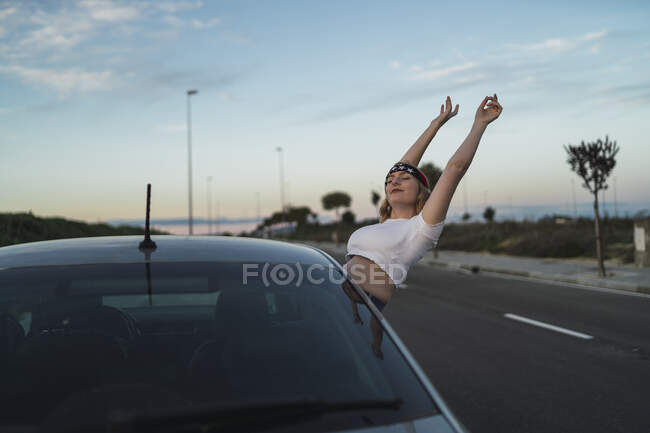 Jovem do sexo feminino em casual desgaste e headband com impressão bandeira americana inclinando-se para fora da janela do carro e levantando as mãos enquanto desfruta de liberdade durante a viagem rodoviária ao pôr do sol — Fotografia de Stock
