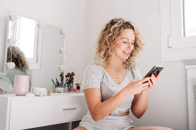 Позитивна молода жінка з кучерявим світлим волоссям в повсякденному одязі переглядає на смартфоні, сидячи біля дзеркала у ванній — стокове фото