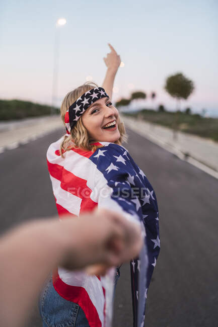 Encantada mulher americana envolta em bandeira dos EUA de mãos dadas com o homem e andando ao longo da estrada enquanto olha para a câmera — Fotografia de Stock
