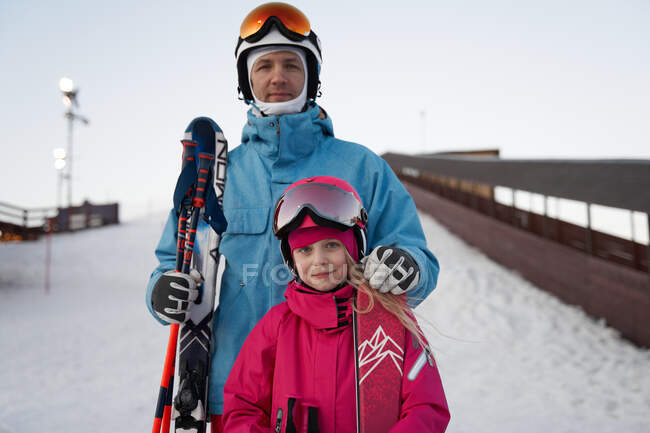 Pai feliz e filha vestindo roupas de esportes quentes e capacetes de pé com esquis na encosta da colina nevada e olhando para a câmera contentamente — Fotografia de Stock