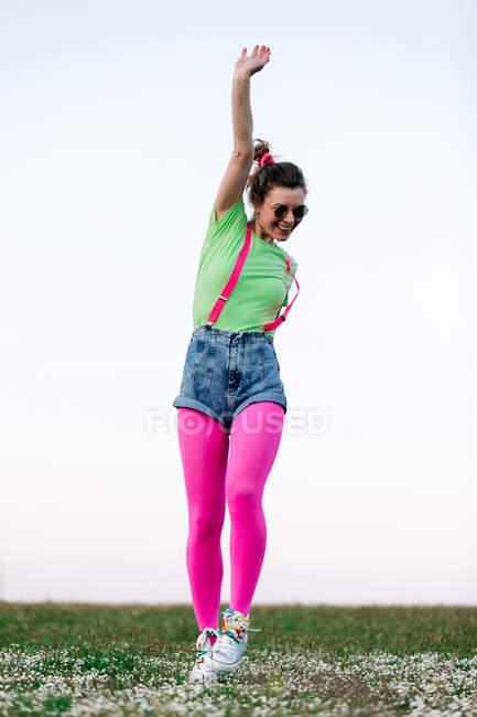 Corpo inteiro encantado jovem mulher vestindo shorts e calças rosa brilhante saltando alegremente com o braço levantado na exuberante clareira gramada na natureza — Fotografia de Stock