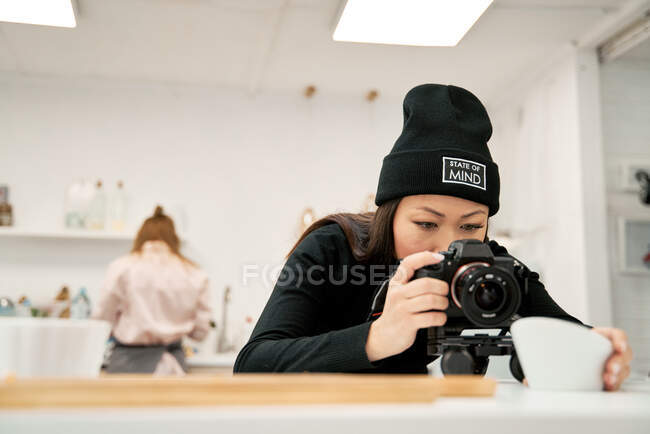 Этническая женщина в шапочке фотографирует миску на цифровую камеру против неузнаваемого партнера на кухне — стоковое фото