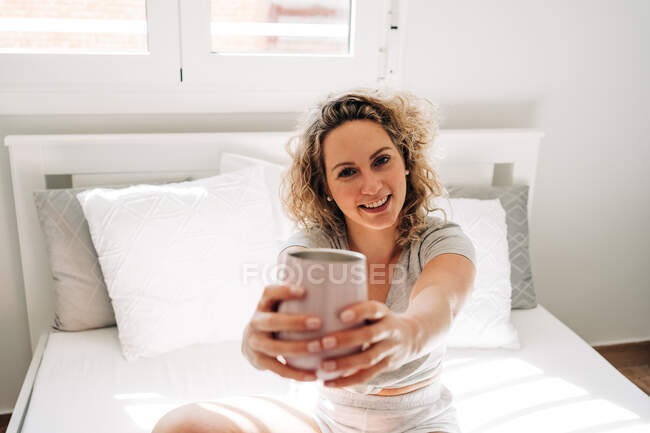 Fröhlich lächelnde Frau streckt Arme mit Becher aus und blickt in die Kamera, während sie auf einem gemütlichen Bett im hellen Schlafzimmer sitzt — Stockfoto