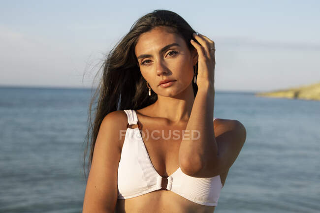 Young pretty female in swimwear looking at camera while standing on sandy shore against ocean under cloudy blue sky — Stock Photo