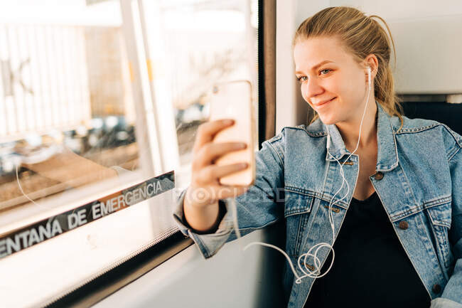 Joven rubia con chaqueta vaquera escuchando música y tomando selfie con teléfono móvil mientras monta en tren sentada cerca de la ventana - foto de stock