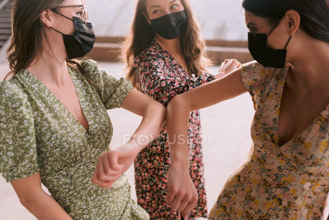 Conteúdo melhores amigas em vestidos ornamentais e máscaras faciais de pano tocando cotovelos enquanto olham uma para a outra na cidade durante a pandemia de coronavírus — Fotografia de Stock