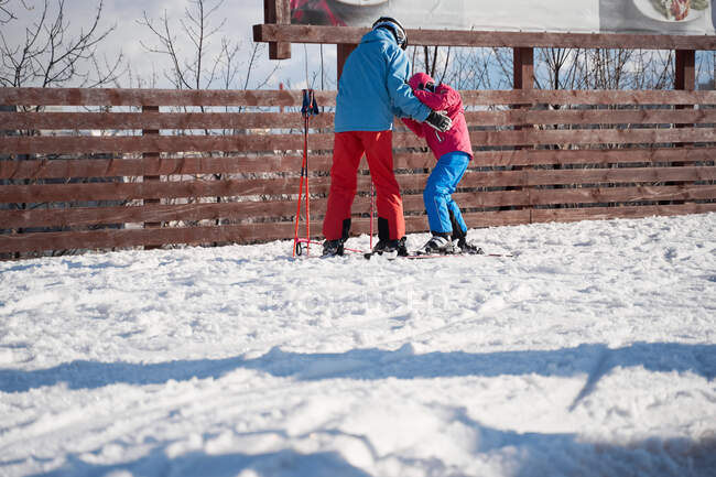 Безликий родитель в теплой спортивной одежде и шлеме учит маленького ребенка кататься на лыжах вдоль снежного склона на зимнем горнолыжном курорте — стоковое фото