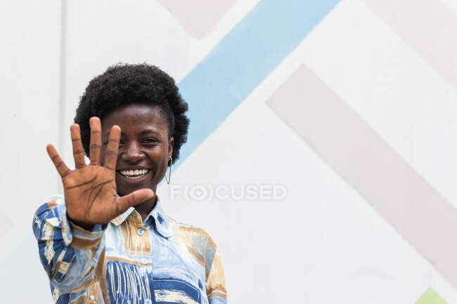 Porträt einer fröhlichen jungen Afroamerikanerin mit lockigem Afro-Haar, die die Hand ausstreckt und sich vor der Kamera versteckt, während sie Spaß hat — Stockfoto