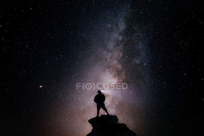 Low angle of silhouette of anonymous tourist standing with light torch on head on cliff against glowing starry sky at night — Stock Photo